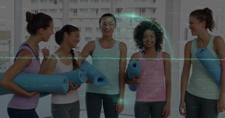 Group of five women holding blue yoga mats and chatting in fitness studio, wearing workout clothes