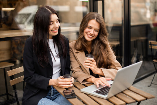 Two young cheerful women friends sitting in a street cafe, drinking coffee and using a smartphone and laptop for work or online shopping. Online purchases on Black Friday or Cyber ​​Monday. - Powered by Adobe