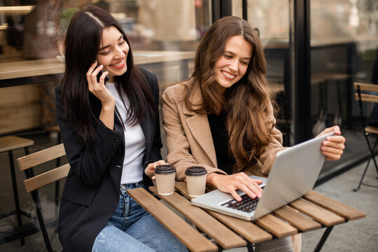 Two young cheerful women friends sitting in a street cafe, drinking coffee and using a smartphone and laptop for work or online shopping. Online purchases on Black Friday or Cyber ​​Monday.