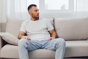 Obraz premium Thoughtful man sitting on a cozy sofa, reflecting in a serene indoor environment with natural light. Stylish outfit complements modern decor and calm atmosphere.