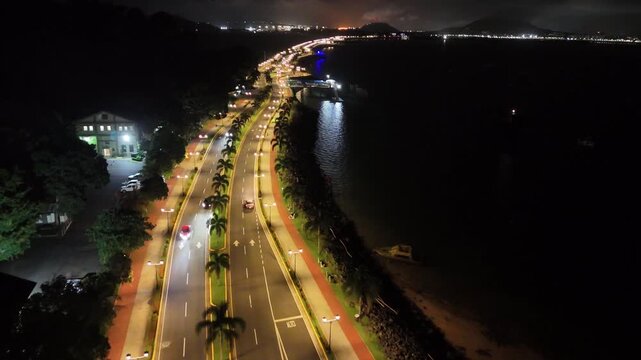 Drone flies along Avenida Amador at night, with streetlights and moving traffic, dark waters on both sides, heading toward Perico and Flamenco Islands in Panama