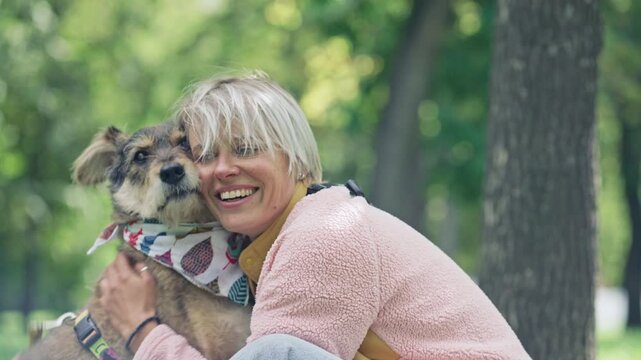 Young happy woman petting, treating and hugging dog wearing patterned bandana during summer day together in sunlit park