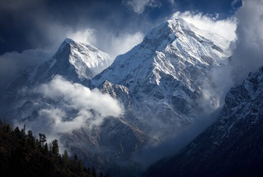 Majestic, snow-capped mountain peaks pierce through swirling clouds in a dramatic landscape with dark trees at the base under a blue sky - Powered by Adobe