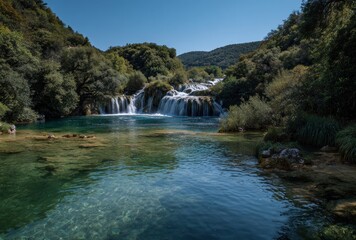Obraz premium Cascading waterfalls flow into a clear, shallow river surrounded by lush green trees under a clear blue sky on a sunny day