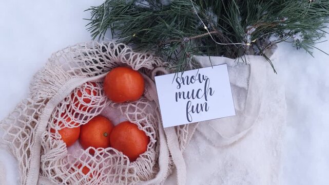 SNOW MUCH FUN text on postcard Mesh shopping bag with tangerines with fir branch and garland in snowy winter outdoor. Go green concept eco-friendly sustainable Christmas. Flat lay in snow background