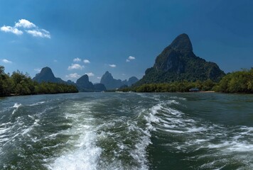 Watery wake through scenic Asian bay. Rocky mountains covered in trees line the waterway under a clear blue sky with a few white clouds