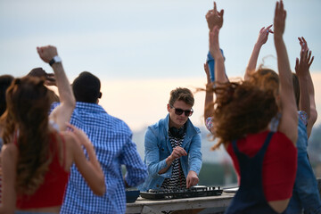 Group of multiethnic young adults dancing outdoors while Caucasian young adult man DJ operating mixing console, people raising hands and enjoying music at rooftop party