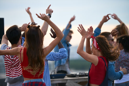 Group of multiethnic young adults dancing outdoors with raised arms, enjoying music together near DJ booth, diverse men and women moving energetically during rooftop party event