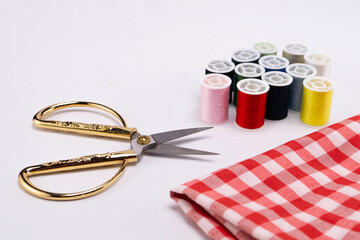 Sewing Essentials - Golden Scissors, Colorful Threads, and Red Checkered Fabric on White Background.