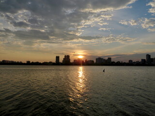 Naklejka premium Bold Sun Path on Water at Golden Hour — Urban Skyline Silhouette, Dramatic Layered Clouds