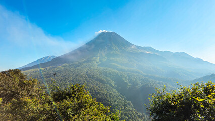 A view of Mount Merapi in the morning with a lush forest in the foreground. The panoramic beauty of Mount Merapi on a clear morning is clearly visible from a distance.