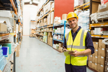 Male worker wearing hard hat inspecting boxes in warehouse aisle with clipboard, copy space
