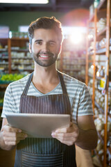 Adult male clerk wearing apron, holding tablet, checking stock in grocery aisle with wooden shelves