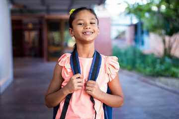 Girl wearing pink top holding blue backpack and smiling in school corridor with yellow flower clip