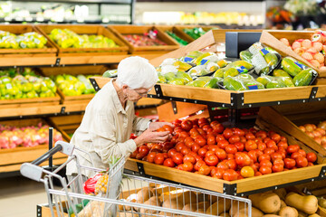 Senior woman examining tomato from bin in produce aisle, pushing cart with avocados and apples