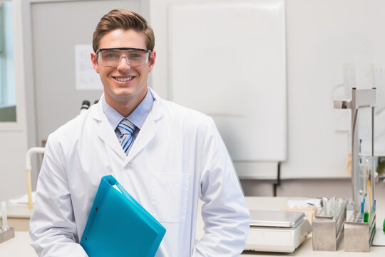 Male scientist standing in laboratory wearing white lab coat and goggles while holding blue folder
