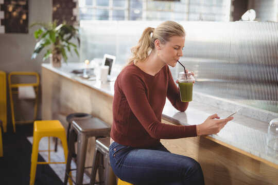 Woman sitting on yellow metal stool at café counter sipping green smoothie and checking smartphone