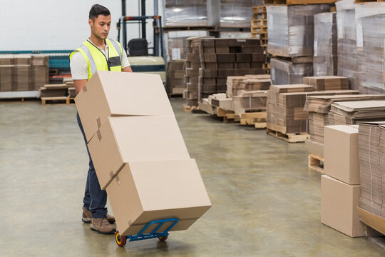 Hispanic male in safety vest pushing blue hand truck with cardboard boxes across warehouse floor