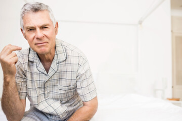 Senior man in pajamas sitting on bed gesturing to lamp and bottle on nightstand copy space