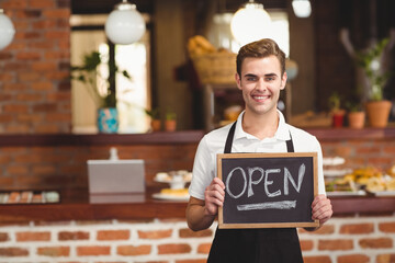 Male barista standing behind brick counter holding open chalkboard sign in bakery, copy space