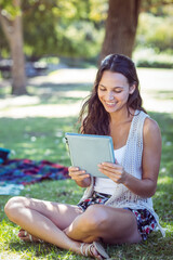 Obraz premium Woman sitting cross-legged on grass in park holding tablet near picnic blanket smiling at screen