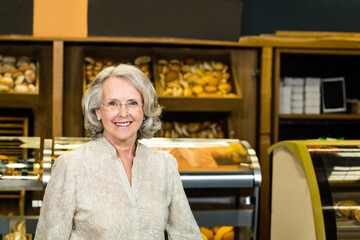 Senior woman standing behind bakery counter showcasing assorted pastries in glass display cases