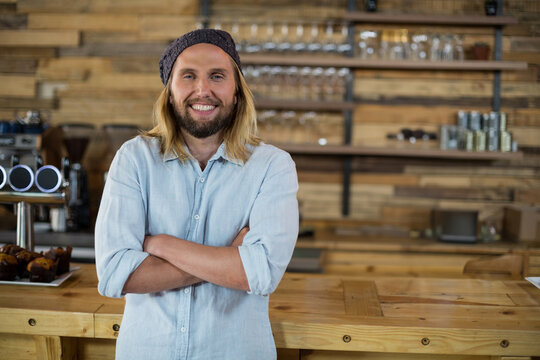 Mid adult male barista smiling, crossing arms behind counter at coffee shop by espresso machine