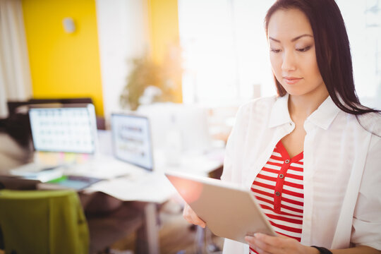 Asian woman holding tablet while checking computer monitors at open-plan office desk, copy space