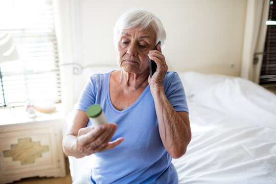 Senior woman holding prescription bottle and talking on smartphone while sitting on bed in bedroom