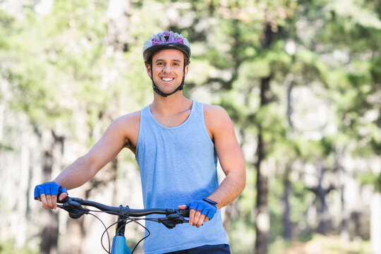 Male cyclist standing smiling on sunlit forest trail holding bike handlebars wearing helmet