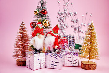 Christmas card on which two cute gnomes received gifts in a small forest of toy Christmas trees on a pink background, horizontal