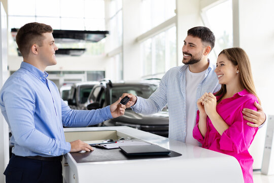 Smiling car dealer handing over keys to happy couple at dealership counter, man and woman receive vehicle keys after signing contract. Lifestyle, trust and transport