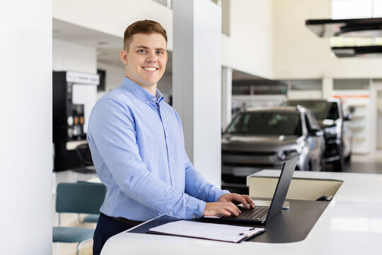 Handsome young car salesman posing at work desk, smiling at camera, positive millennial man using laptop in auto dealership center. Automobile manager standing in showroom store, copy space