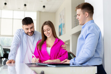 Fototapeta premium Smiling woman signing car purchase agreement while man and dealer watch at showroom desk, couple finalizing automobile contract. Lifestyle, finance and transportation