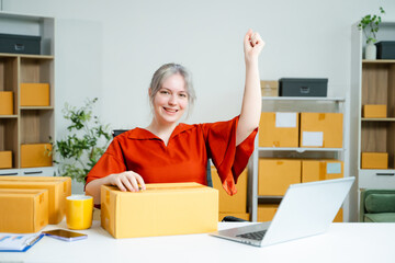 Young Caucasian woman celebrating a successful online business at home, surrounded by boxes and...