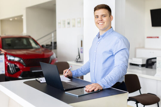 Smiling car dealer working with laptop at showroom desk. Young professional man checking documents and contracts near new vehicles, lifestyle and automobile retail