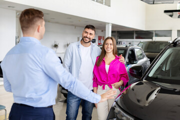 Obraz premium Smiling couple listening to car dealer while viewing vehicle in showroom. Man and woman discussing automobile options with salesman, closeup