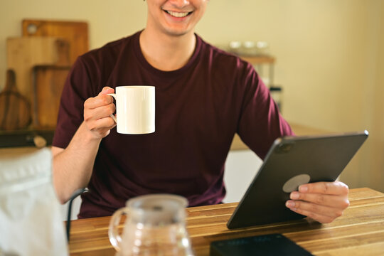 Smiling man holding a white coffee mug while using a digital tablet at home kitchen