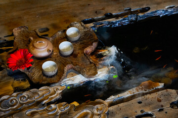 A wooden, leaf-shaped tray holds a small brown teapot and three matching teacups on a wooden trunk table with mist coming out