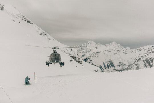 A man sits next to a helicopter landing in the mountains.