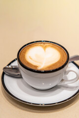 A white coffee mug decorated with a heart-shaped milk foam pattern. The mug sits on a saucer against a yellow background. A minimalist composition, perfect for coffee-related topics and relaxation.