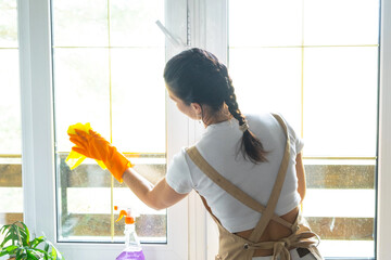 Woman in rubber gloves and apron manually washes window balcony of house with rag cleaner and mop inside interior. Restoring order and cleanliness in spring, cleaning servise