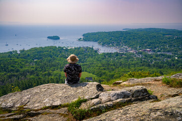 A hiker sits on a rocky ledge at the summit of Mt. Battie in Camden Hills State Park, overlooking Camden Harbor and Penobscot Bay in Maine.