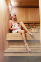 Young woman relaxing and sweating in hot sauna wrapped in towel. Girl In Sauna. Interior of Finnish sauna, classic wooden steam room with hot steam. Russian bathroom. Relax in hot Bathhouse with steam