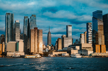 Obraz premium Manhattan skyline shines under evening clouds. Golden sunlight reflects off modern buildings in Manhattan, New York, as dark clouds loom in the vibrant evening sky.