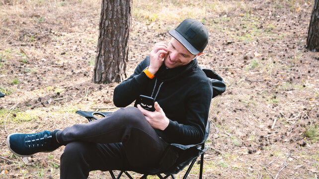 Happy man in a black hoodie and cap, laughing while looking at his smartphone in a forest setting. Relaxed outdoor activity, enjoying digital content in nature.