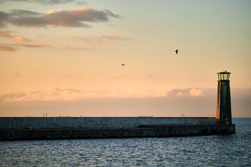 Fototapeta premium A lighthouse standing at the edge of a long pier extending into the sea, with the golden glow of the setting sun casting soft light on the water and sky.