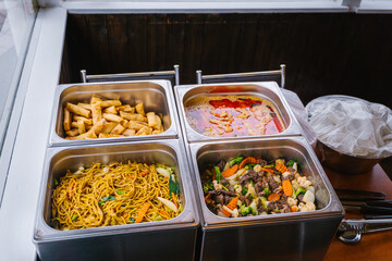 Buffet with Thai food including noodles, spring rolls, curry, and stir-fried vegetables in stainless steel trays.