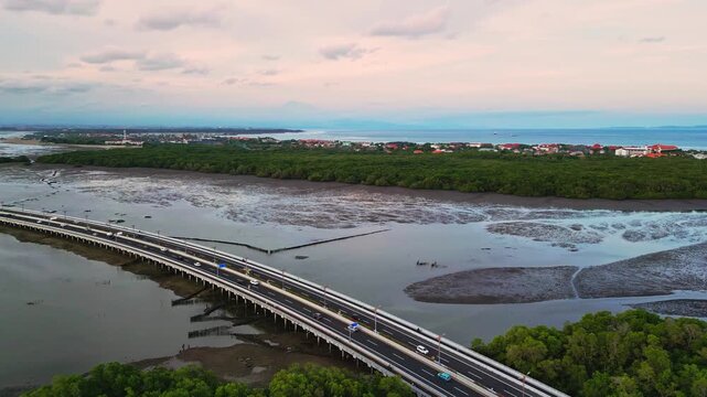 Aerial drone footage captures the Bali Mandara Toll road bridge extending across wide muddy flats revealed at low tide with a tropical Indonesian landscape creating a striking coastal highway scene