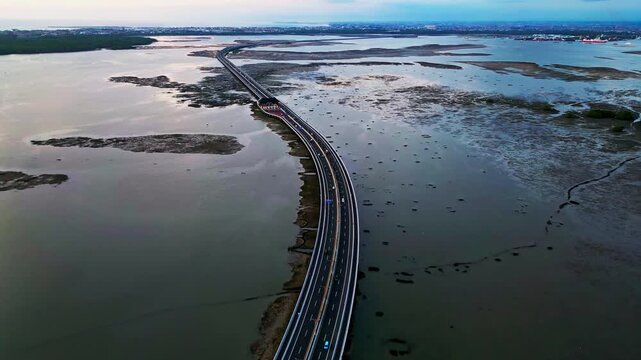 Drone captures sweeping views of Bali Mandara Toll road bridge crossing muddy low tide terrain framed by tropical vegetation and coastal atmosphere highlighting Indonesian infrastructure.